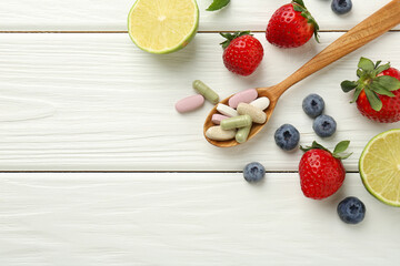 Different vitamin pills in spoon and fresh fruits on white wooden table, flat lay. Space for text