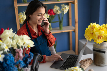 Florist small business flower shop owner. Woman using mobile phone and laptop to take orders for her floral store. Hispanic gardener taking client order during conversation