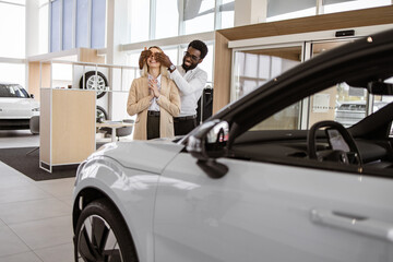 African American boyfriend covering eyes of his Caucasian young girlfriend for surprise in car showroom. Happy husband buying to his loving wife new vehicle at car dealership.