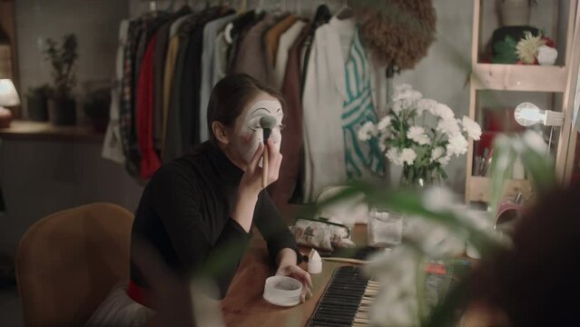 Young actress with mime makeup applying face powder with brush while sitting at vanity desk in dressing room and preparing for performance