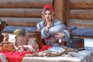 A beautiful Russian girl in traditional Russian clothes and a kokoshnik stands at a table with...