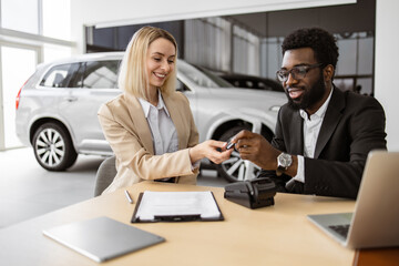 Fototapeta na wymiar Happy Caucasian young woman sitting at table and receives keys of SUV car from African man salesman in suit in bright showroom. Concept of buying new modern vehicle.