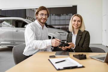 Female confident manager helps to male customer make payment for purchase of branded crossover. Happy young man paying with electronic credit card at terminal for purchase of new modern car.