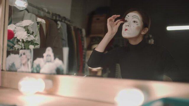 Female mime artist with stage makeup applying false eyelashes in front on illuminated mirror at vanity desk in dressing room
