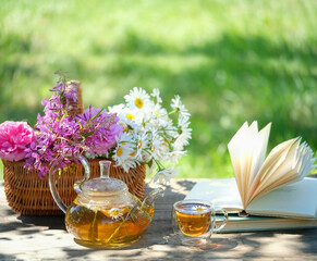 glass teapot, cup with herbal tea, flowers in basket and book on table in garden. Summer nature...