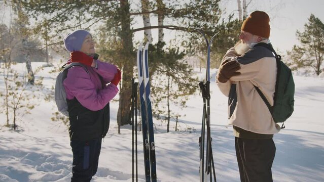Medium full lockdown slowmo shot of senior Caucasian husband and wife standing in winter forest, stretching arms after cross-country skiing, then taking gear and walking away