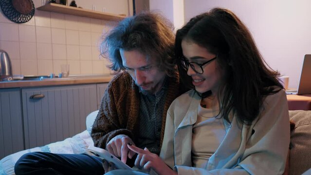 Young couple on couch, closely sharing content on smartphone, in comfortable living space with kitchenette in background. 