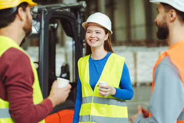 Beautiful, young woman in hard hat holding cup of coffee, looking away, listening to her colleagues