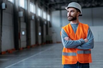 Smiling bearded professional engineer wearing protective workwear, white hard hat with crossed arms