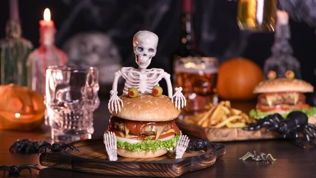 A witch's hand opens a bottle of beer and pours it into a glass in the background, on the table there is a Monster Burger and fries. Halloween party idea. Real time