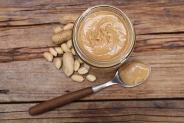 Tasty peanut nut paste in jar and spoon on wooden table, flat lay