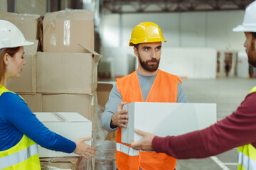 Middle aged worker wearing hard hat and uniform, holding box, talking with colleagues in warehouse