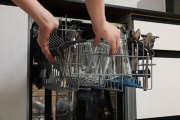 Woman loading dishwasher with glass and cutlery indoors, closeup