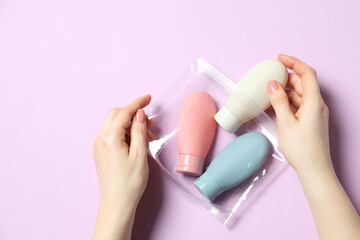Cosmetic travel kit. Woman putting small bottle with personal care product into plastic bag against violet background, top view
