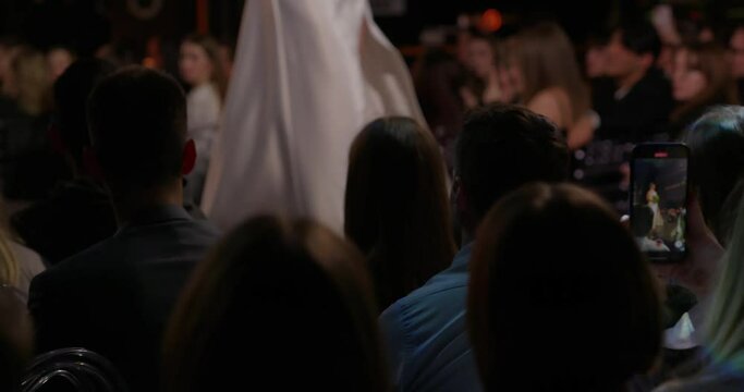 A view over the heads of the audience during fashion week. A model in a white dress walks down the catwalk showing off a new collection to the audience and fashion designers. High quality 4k footage
