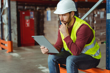 Serious, pensive, bearded man, foreman wearing hard hat and workwear, using digital tablet, thinking