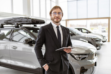 Portrait of confident Caucasian man, salesman in suit in bright modern car service. Young successful manager on background of rows of new car dealership crossovers. - obrazy, fototapety, plakaty