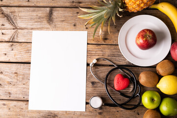 Directly above shot various fruits with stethoscope and heart shape by blank paper on wooden table
