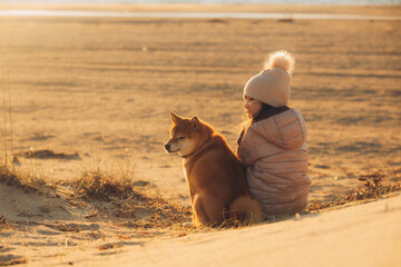 A young girl with a dog in nature. Kid girl sitting with a shiba inu dog on the beach at sunset in Greece in winter - 786304484
