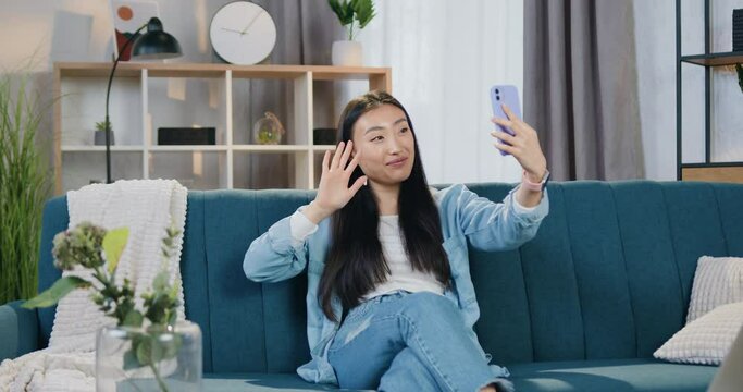Asian woman in casual clothes using smartphone sitting on the couch at home and enjoying her video call with friend. Leisure concept.
