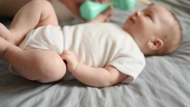 Infant lying on his back, awake, holding a rattle, playing with it, newborn baby smiling, soft focus, focus on legs. Concept of motherhood, maternity leave, child care, child development.