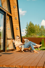 Woman typing on laptop while laying in a folding wooden sun lounger on the terrace of a-frame cabin