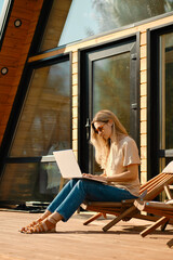 Woman checking mail on laptop sitting in a folding wooden chair on the terrace of summer cabin