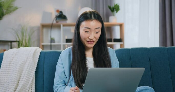 Close up portrait of asian woman using laptop sitting on the couch at home and waving hand to greet her interlocutor via video call