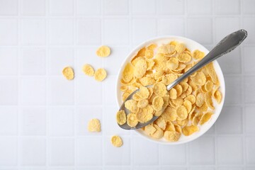 Breakfast cereal. Tasty corn flakes with milk in bowl and spoon on white tiled table, top view....