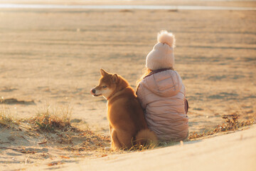 A young girl with a dog in nature. Kid girl sitting with a shiba inu dog on the beach at sunset in Greece in winter - 786323088