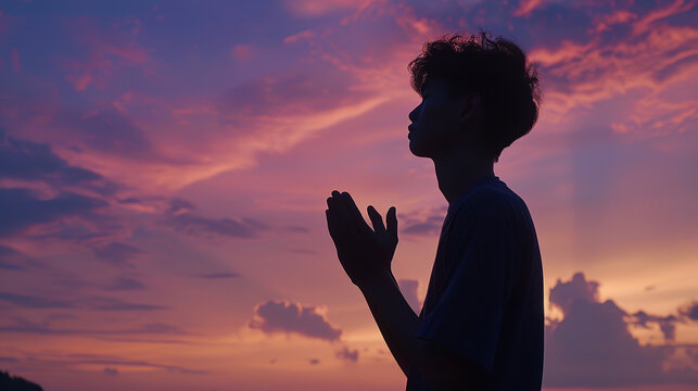 Picture a young man silhouetted against the backdrop of a serene, softly lit sky at dawn or dusk. He stands with his eyes closed and hands clasped in prayer