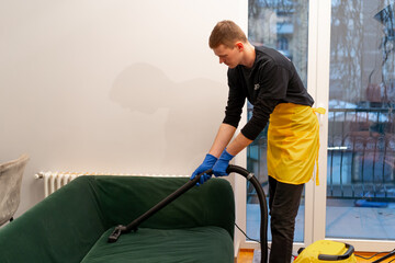 in an apartment young cleaner in a yellow apron professionally vacuums a green velor sofa
