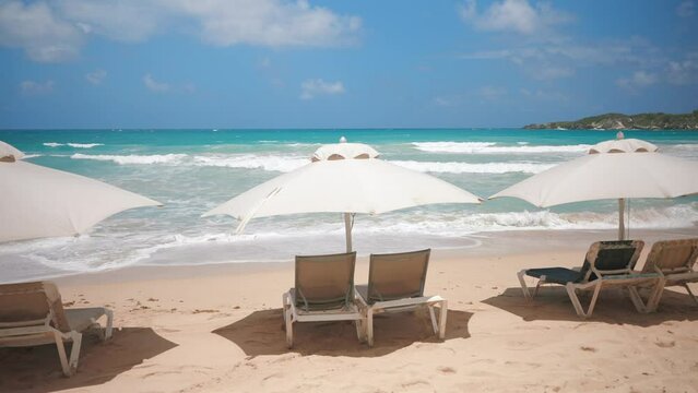 A row of sun loungers under white parasols on the sandy beach near the sea water with big waves. Rest and relaxation summer holiday scene