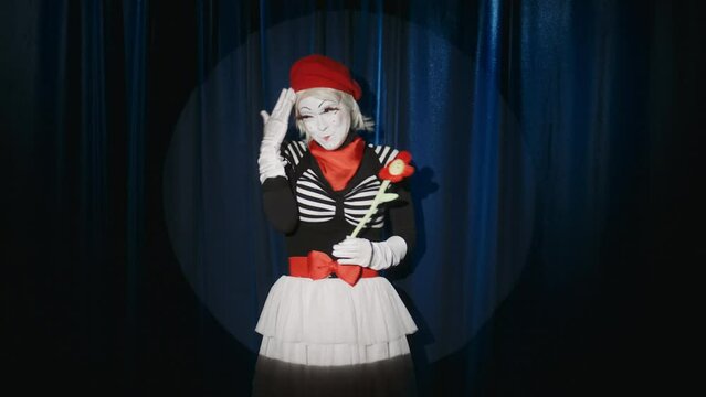 Funny female mime artist standing in spotlight on theatre stage, hugging flower and acting on camera while giving performance