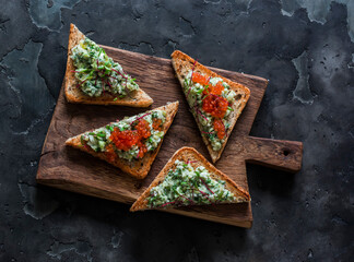 Delicious tapas, appetizer, snack - bruschetta with avocado, eggs, gherkins, greens spread and red caviar on a wooden chopping board on a dark background - 786362023