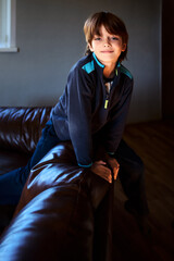 Handsome young Caucasian boy leans on back of leather sofa in room with gray walls illuminated by...