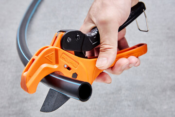 Orange ratcheting PVC pipe cutter tool in hand of plumber cutting off piece of pipe on gray background.