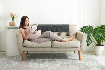 Young beautiful asian woman reading a book at home. Brunette female and rough coated dog sitting on textile couch. White wall background, copy space - obrazy, fototapety, plakaty