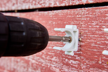 Plumber is using cordless drill to secure white pipe clips to weathered red wooden house for organizing pipeline outdoors.