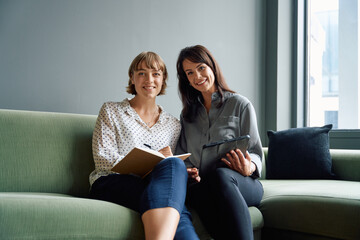 Two happy business women sitting on sofa using digital tablet during meeting in office