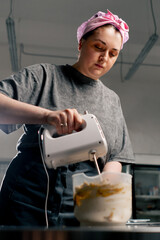 female baker in a professional kitchen whips paste for cream on a cake with a mixer