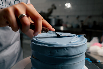 close up female baker in a professional kitchen distributes blue cream onto a sponge cake with a...
