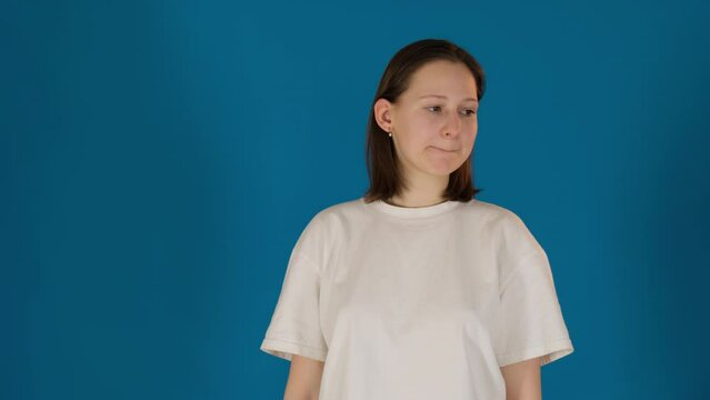 Anxious woman in t-shirt bites lips and shrugs shoulders on blue background. Lady performs obsessive movements caused by stress in studio