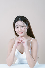 Sexy beautiful Asian woman in white dress with natural makeup and looking at camera isolated over beige background. Fashion shiny highlighter on skin, perfect make-up.