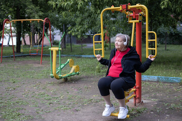 Smiling elderly woman in headphones is doing exercises on simulator, trainer outdoors in the park. Active life of pensioners. Adaptation of pensioners in the modern world. Prevention of mental illness