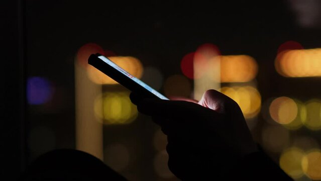 Close-up of female hands holding mobile phone on the background of night city lights. Unrecognizable woman types text messages while sitting on the windowsill in the dark room.