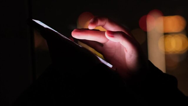 Close-up of woman hands scrolling through the news feed on the phone with night city lights on the background. Unrecognizable person uses smartphone at the window.