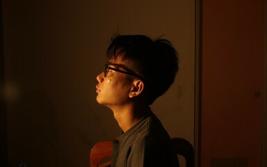 A contemplative young man captured in dramatic lighting, sitting alone in a dim room. The photo...