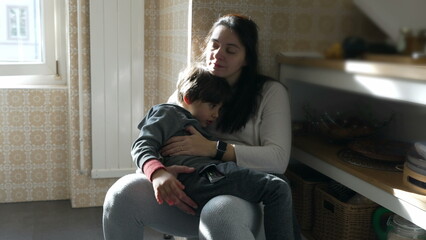 Mother resting while holding 5 year old son at kitchen, authentic family lifestyle scene of tired...