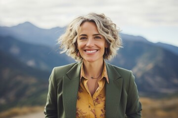Portrait of a grinning woman in her 50s dressed in a stylish blazer while standing against backdrop of mountain peaks